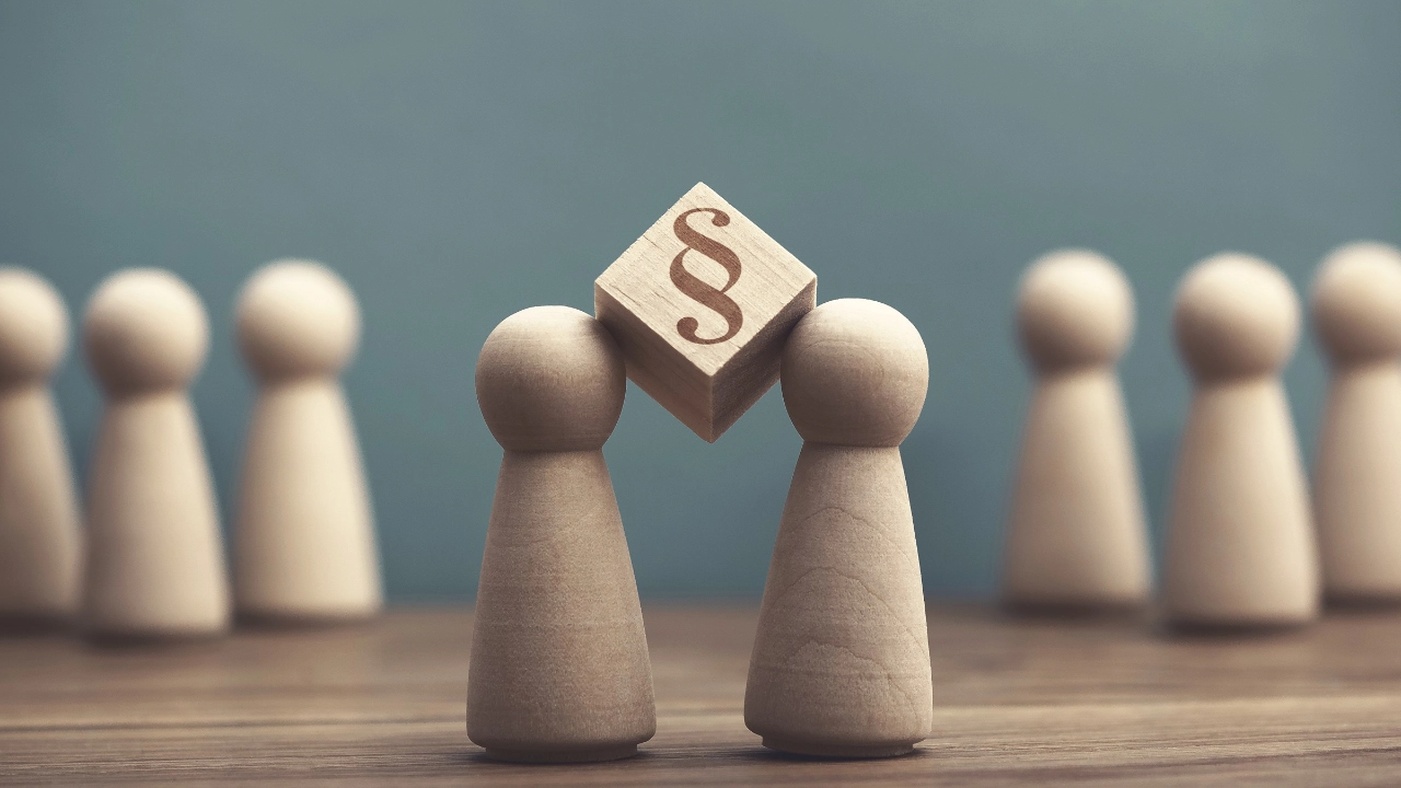 ein Würfel mit Paragraf darauf ist eingeklemmt zwischen den Köpfen zweier Spielfiguren 